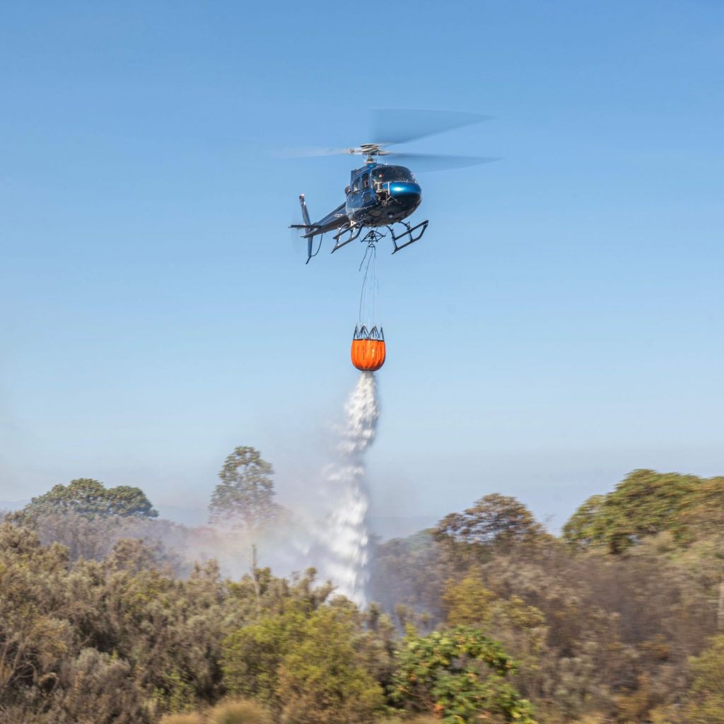 A large orange sack suspended from a helicopter, dropping water over a forest